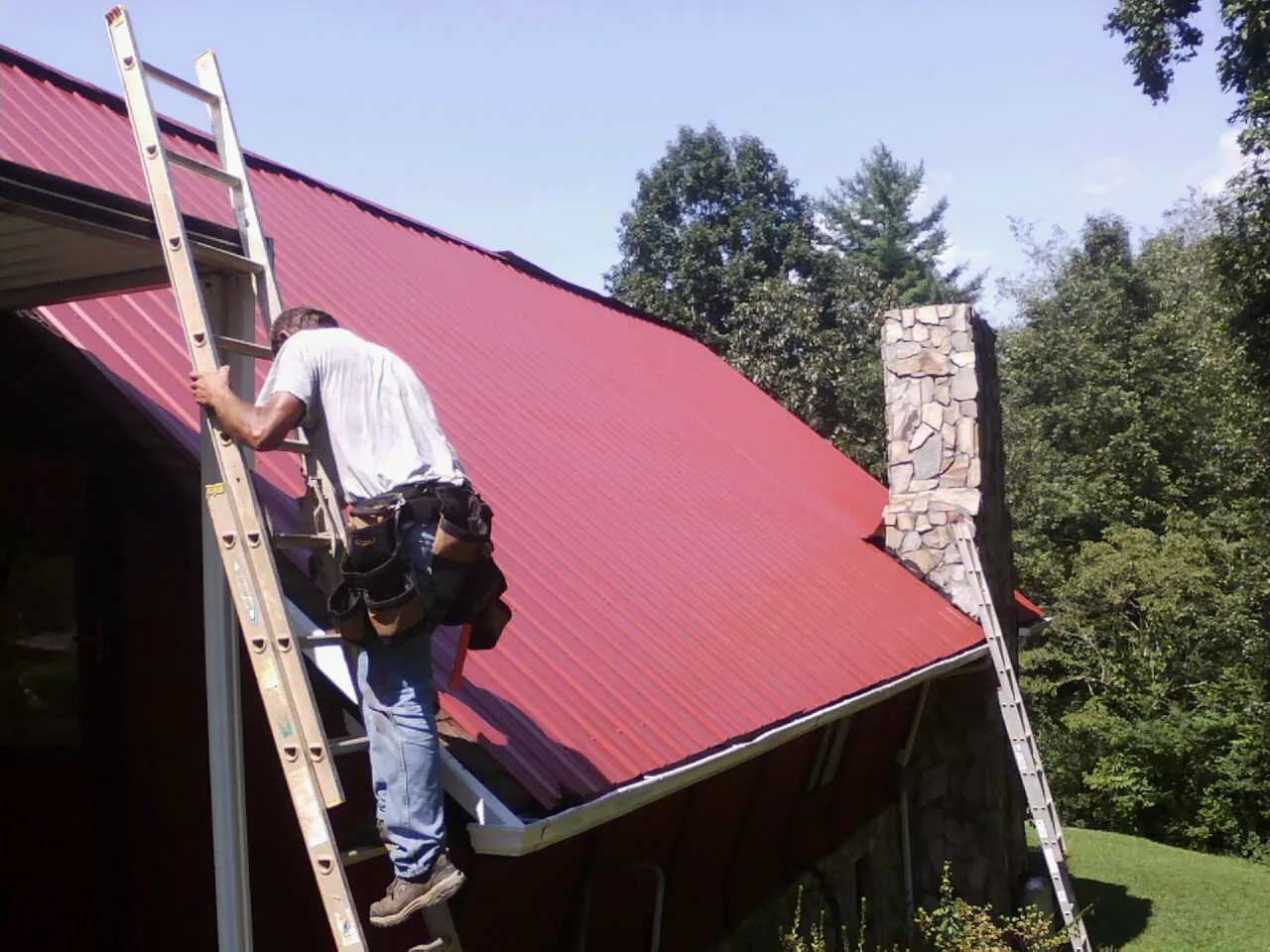 Roofer on a ladder beside a red metal roof and stone chimney — Soffit & Fascia Repair by Roofing Brothers in Birds Landing